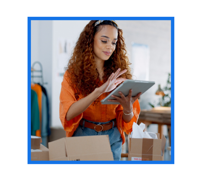 A young woman with curly hair using a tablet while working in a small business environment, surrounded by boxes and packages.